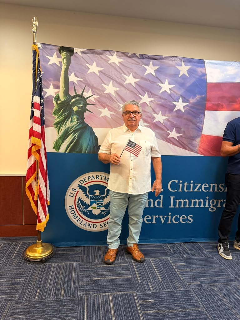 Wilson Almeida segurando a bandeira americana em frente ao painel do USCIS